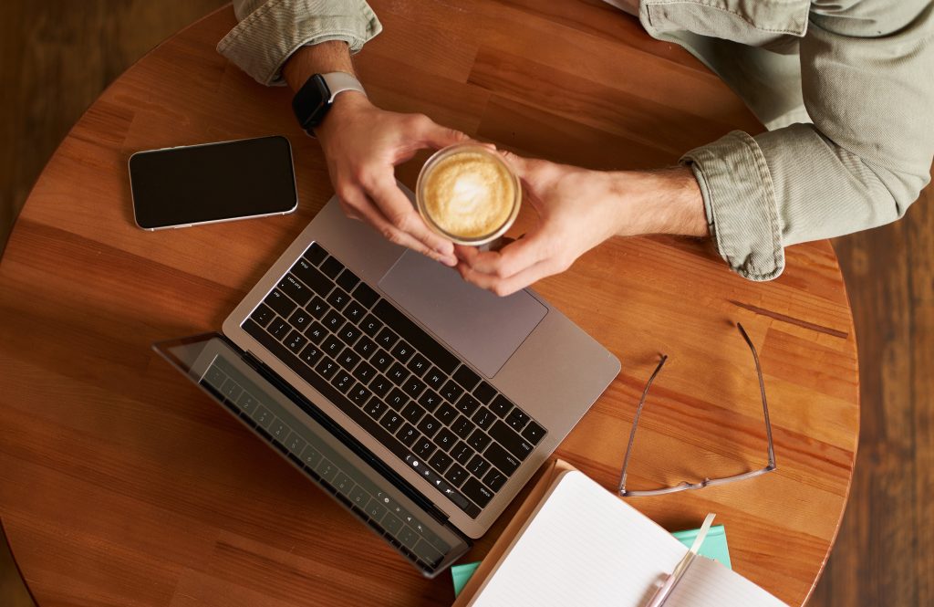 Male hands holding cup of coffee, man sitting at round table in a cafe, working on laptop, had his smartphone and notebook, drinking cappuccino